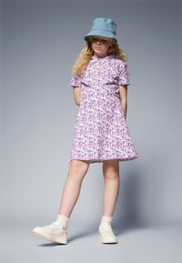 Robe à manches courtes à motif floral rose et blanc, avec un bas évasé ; associée à un chapeau seau en denim bleu et des baskets blanches.