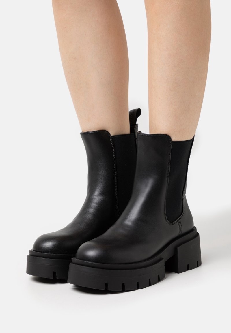 Bottines en cuir noir à semelles plateformes épaisses portées sur des jambes nues, avec des panneaux latéraux élastiques et des languettes à l'arrière.
