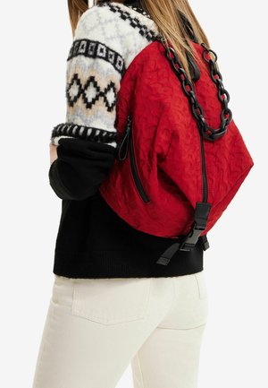 Personne portant un pull à motifs noir et blanc et un pantalon blanc, portant un sac à dos rouge texturé avec des détails en chaîne noire et des fermetures éclair.