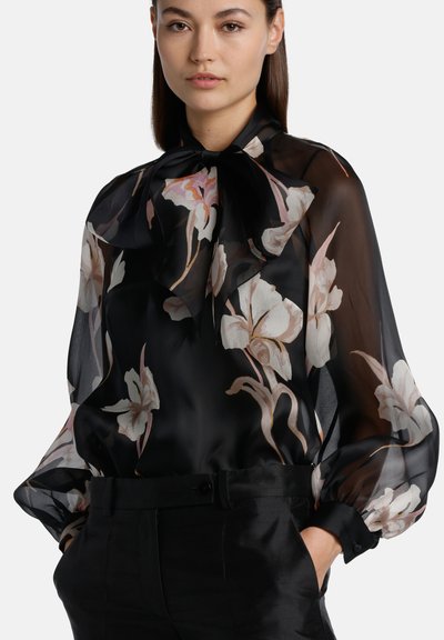 Blusa nera trasparente con stampa floreale, caratterizzata da un nodo al collo, maniche a sbuffo e una consistenza liscia. Il design include grandi fiori bianchi e rosa.