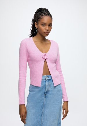Femme aux cheveux tressés portant un cardigan court rose avec un fermoir fleur et une jupe en jean bleue taille haute sur fond uni.