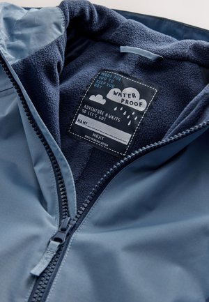 Chaqueta impermeable con exterior azul, suave forro polar azul marino y cierre con cremallera, con una etiqueta de cuidado que detalla las instrucciones de mantenimiento.