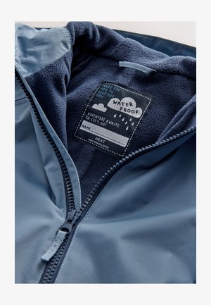 Geacă impermeabilă cu exterior albastru, captuseală moale din fleece bleumarin și închidere cu fermoar, prevăzută cu etichetă de întreținere ce detaliază instrucțiunile de îngrijire.