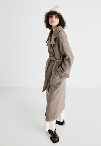 Lungo cappotto in misto lana beige con cintura, ampi rever e grandi bottoni; indossato sopra un vestito bianco e abbinato a scarpe nere chunky.