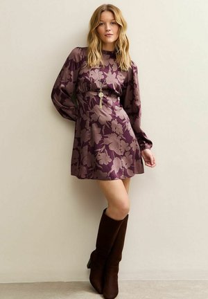 Donna che indossa un mini abito di raso viola con fiori, maniche lunghe a sbuffo e stivali alti fino al ginocchio in suede marrone, in piedi davanti a una parete beige.