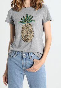 Camiseta gris de manga corta con un diseño de piña decorado con lentejuelas en dorado, verde y azul. Combinada con jeans de mezclilla azules.