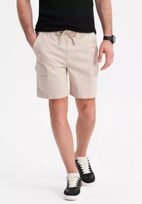 Shorts cargo beiges avec deux poches latérales, une taille élastique et un cordon de serrage. Accompagnés de baskets noires avec des lacets blancs.