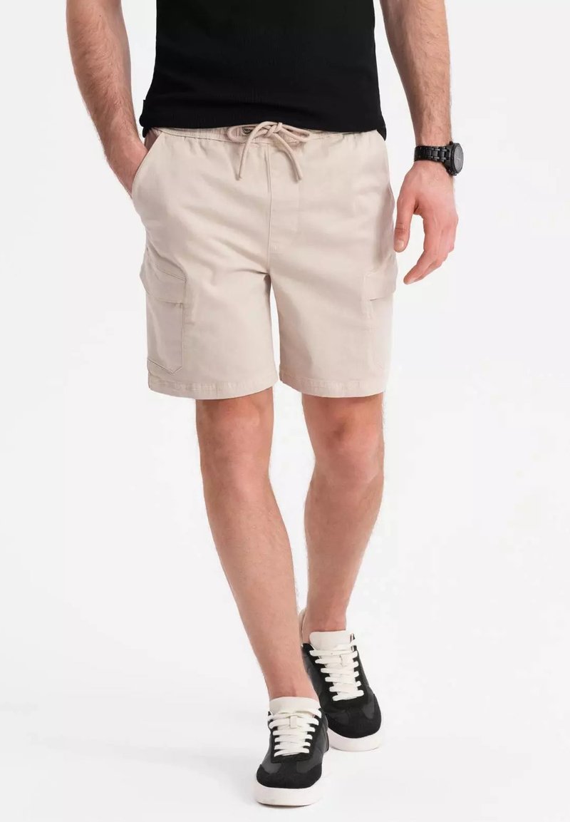 Shorts cargo beiges avec deux poches latérales, une taille élastique et un cordon de serrage. Accompagnés de baskets noires avec des lacets blancs.