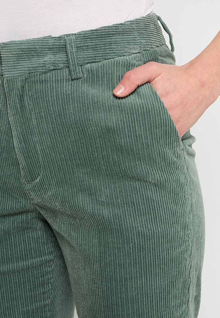 Hand in der Vordertasche von grünen Cordhosen, die mit einem weißen Hemd getragen werden.