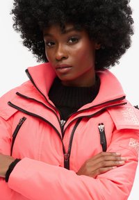 Pink isoleret jakke med flere lynlåse, høj krave og branding på skulderen. Bæres over en sort sweater med struktureret overflade.