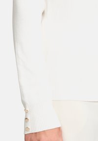 Fehér kötött pulóver ujj, bordás mandzsettával és három dekoratív aranyszínű gombbal. Puha textúra és testhez simuló fazon, sima felülettel.