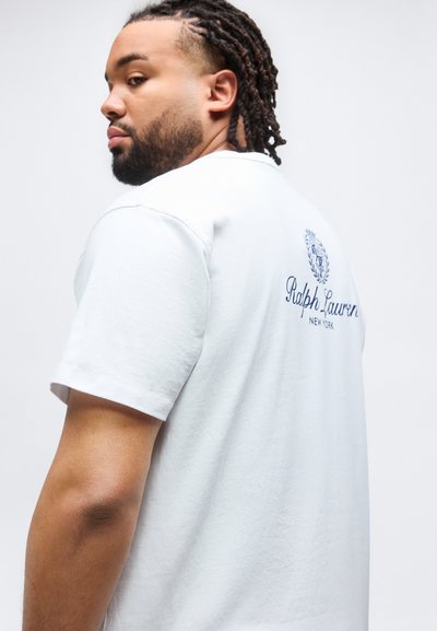 Άνδρας με πλεξουδάκια που φοράει λευκό μπλουζάκι Ralph Lauren, γυρισμένος ελαφρώς προς τα πίσω δείχνοντας το λογότυπο και κοιτάζει πάνω από τον ώμο του.