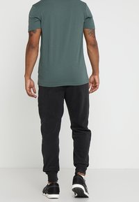 Muž v zeleném tričku s krátkým rukávem, černých joggingových kalhotách a černých teniskách, stojící zády k fotoaparátu na jednobarevném pozadí.