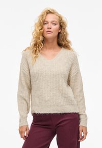 Bež pleten pulover z V-izrezom, dolgimi rokavi in teksturiranim vzorcem. Tkanina je videti mehka, s rahlo puhastim zaključkom.
