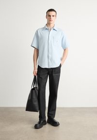 Camicia button-up azzurro chiaro a maniche corte con taschino sul petto, abbinata a jeans neri e scarpe slip-on nere, portando una borsa tote nera.