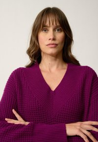Fioletowy sweter dżersejowy z dekoltem w serek, o strukturalnym wzorze i szerokich rękawach. Materiał wydaje się miękki, z widocznymi ściągaczami i otwartym splotem.
