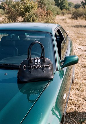 Un sac à main en cuir noir repose sur le capot d’une voiture bleu sarcelle garée à côté d’un champ sec.