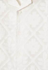 Chemise blanche avec un motif texturé floral et géométrique en relief, montrant trois boutons blancs et le bord du col.