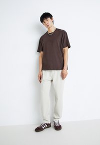 T-shirt en coton marron à manches courtes, associé à un pantalon blanc décontracté et des baskets bordeaux avec des rayures blanches. Design et coupe simples.