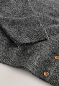 Prim-plan cu mâneca puloverului gri din lână tricotată și nasturii din lemn de pe tiv, evidențiind textura moale a materialului și detaliul manșetei cu nervuri.