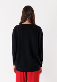 Maglione nero lavorato a maglia con una vestibilità rilassata, scollatura ampia e polsini e orlo a coste. Tessuto morbido con dettagli minimi, indossato con pantaloni rossi.
