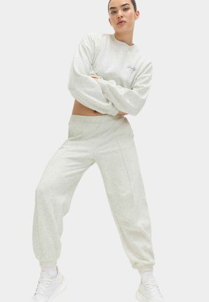 Ung kvinde iført lysegrå cropped sweatshirt, matchende joggers, hvide sokker og hvide sneakers, stående med krydsede arme.