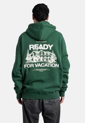 Person, der eine grüne Kapuzenjacke mit weißem Hausmotiv und dem Text "READY FOR VACATION" trägt, von hinten zu sehen.