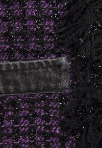 Gros plan d’un tissu texturé noir et violet avec des fils brillants, montrant un bord de poche noir et des fibres effilochées sur un côté.