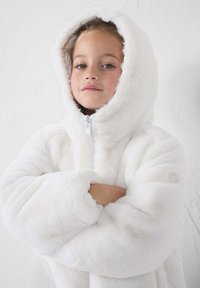 Chaqueta de pelaje sintético blanco con capucha y cierre frontal, mangas amplias y textura suave, llevada por una chica con los brazos cruzados y un fondo neutro.