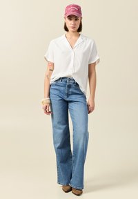 Camisa corta de manga larga blanca con botones, jeans de cintura alta acampanados de color azul claro y una gorra rosa desgastada con el texto "CALIFORNIA". Los accesorios incluyen pulseras.