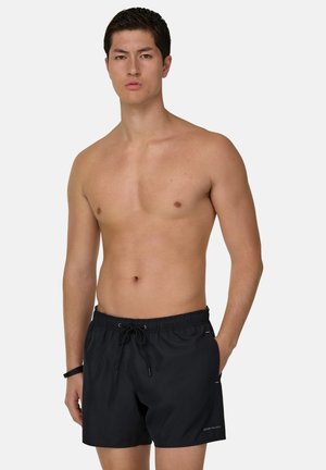 Giovane uomo senza maglietta che indossa pantaloncini da bagno neri con coulisse, in piedi con una mano in tasca su sfondo bianco.