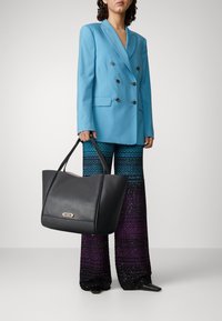 Black leather tote bag with a structured shape, featuring a gold hardware accent. The model wears a blue blazer and ombre patterned wide-leg pants.