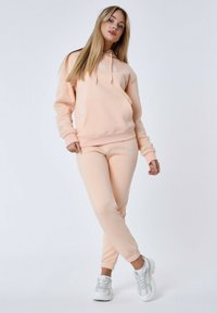 Licht perzikkleurig sweatshirt met capuchon en voorzak, gecombineerd met bijpassende joggers. Beide gemaakt van zachte stof, met elastische manchetten en logo-details.