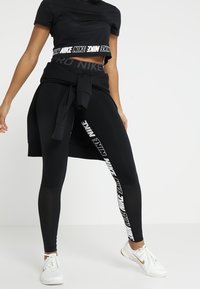 Svart atletisk crop-top med en texturerad design och en djärv vit "Nike"-logotyp. Svarta leggings med sidobranding och hög midja. Vita sneakers.