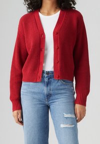 Punainen neulecardigan, jossa on ribbikuvio, V-pääntie ja kuusi nappia. Yhdistetty vaaleansinisiin kuluneisiin farkkuihin.