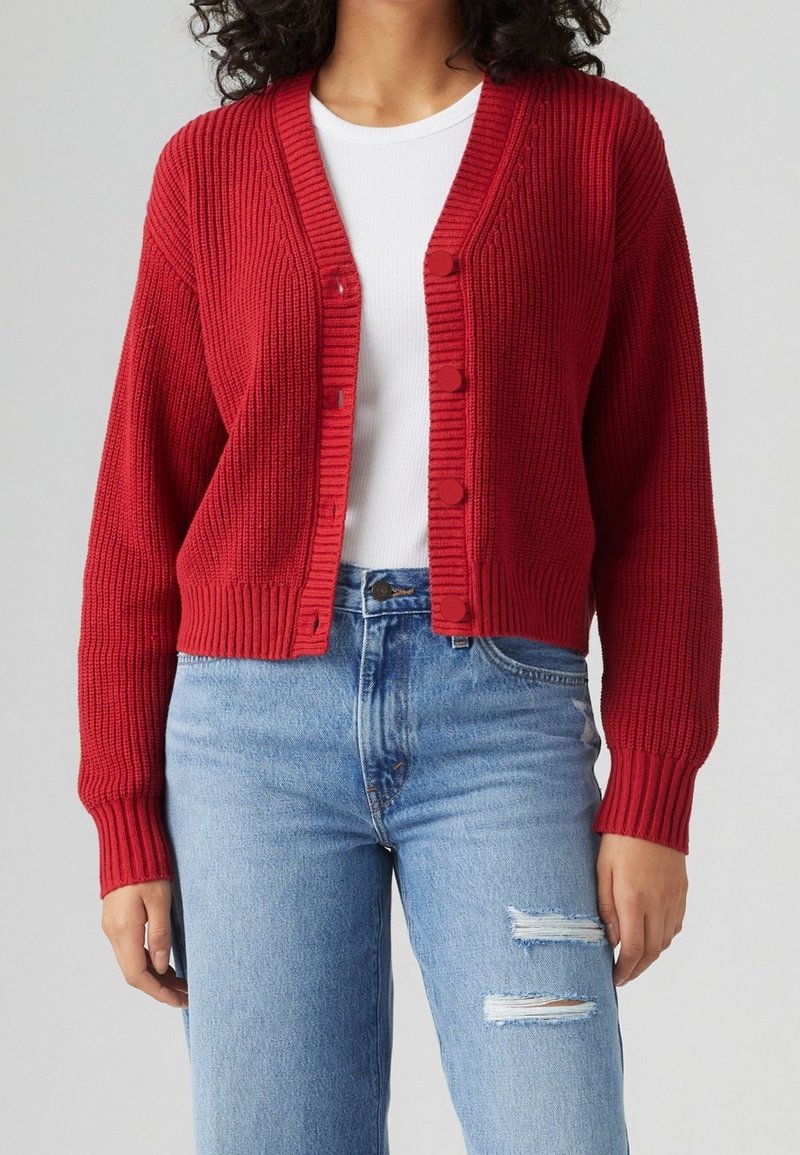 Punainen neulecardigan, jossa on ribbikuvio, V-pääntie ja kuusi nappia. Yhdistetty vaaleansinisiin kuluneisiin farkkuihin.