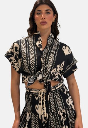 Donna che indossa una camicia nera e crema a maniche corte con bottoni, annodata in vita con gonna coordinata a vita alta.