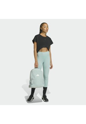 Zaino grigio chiaro con logo Adidas, indossato con un top corto nero e leggings verde chiaro. Tessuto liscio, design sportivo e sneakers nere.