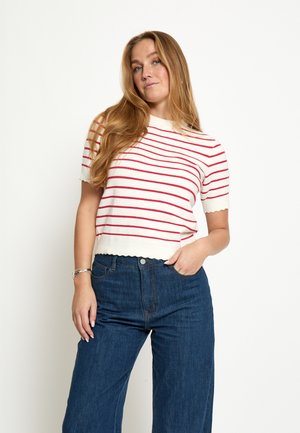 Frau mit langen blonden Haaren, die eine weiße Kurzarmbluse mit roten Streifen und hoch taillierte, weit geschnittene blaue Jeans trägt, mit der Hand in der Tasche.