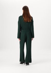 Green cable-knit jumpsuit with long sleeves, wide-leg pants, and a belted waist, featuring a textured pattern throughout.