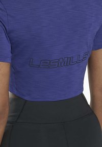 T-shirt de sport court violet avec tissu en maille, manches courtes et logo "LES MILLS" en noir à l'arrière. Associé avec des leggings taille haute noirs.