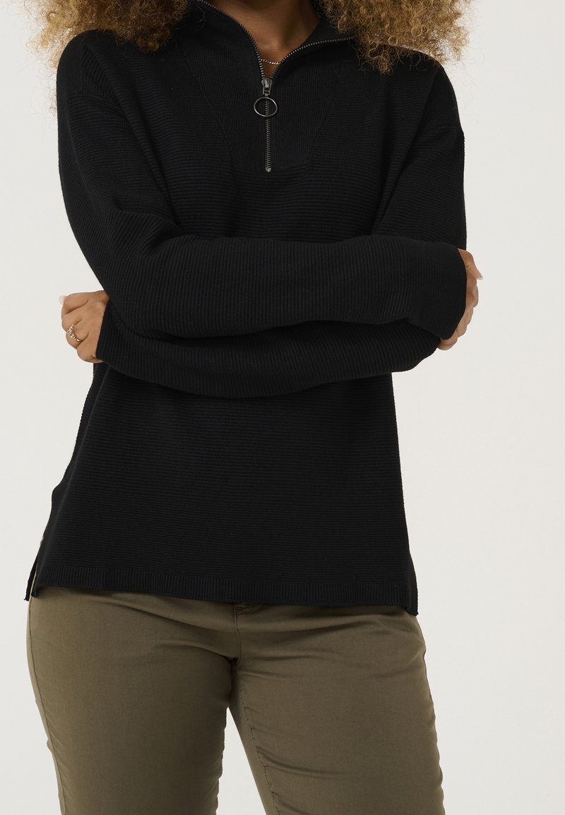 Svart tröja med dragkedja i kragen, ribbad textur och sidofickor. Bärs över olivgröna byxor, med långa ärmar.