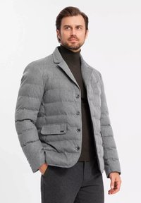 Chaqueta tipo puffer gris con un diseño estructurado y acolchado, que presenta un cuello de muesca, botones frontales y un único bolsillo en el pecho.