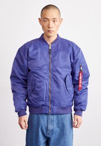 Chaqueta bomber morada hecha de material brillante, con cierre frontal, puños acanalados, bolsillos laterales y un bolsillo en la manga con cremallera y etiquetado.
