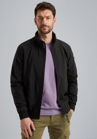 Schwarze leichte Bomberjacke mit Stehkragen, Reißverschluss vorne und elastischen Bündchen, getragen über einem lavendelfarbenen T-Shirt, kombiniert mit olivgrünen Cargo-Hosen.