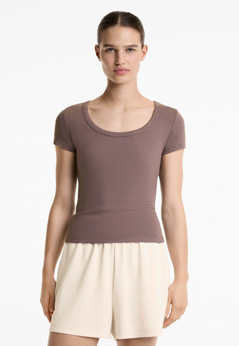 Top corto a maniche corte marrone a coste con scollo rotondo, abbinato a pantaloni corti plissettati beige chiaro, creando una silhouette semplice e aderente.