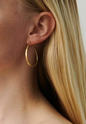 Lähivõte blondi naise kõrvast, kes kannab keskmise suurusega kuldset rõngaskõrvarõngast, sirged juuksed on kõrvast taha kinnitatud.