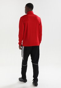 Rød langermet genser med høy krage, kombinert med svarte treningsbukser med hvite striper langs sidene og stilige sko.