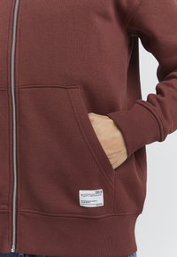 Burgunderfarget zip-up hettegenser laget av mykt tekstil, med frontlommer, ribbede mansjetter, og en etikett med pleieinstruksjoner på den nedre siden.