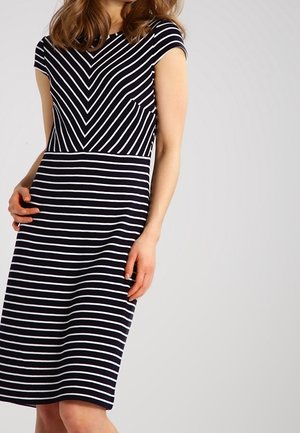Femme portant une robe noire avec des rayures blanches ; le haut a des rayures diagonales formant un V, et la jupe a des rayures horizontales.